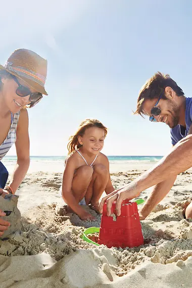 Mutter, Vater und deren Tochter sitzen am Strand und bauen Sandburgen. 