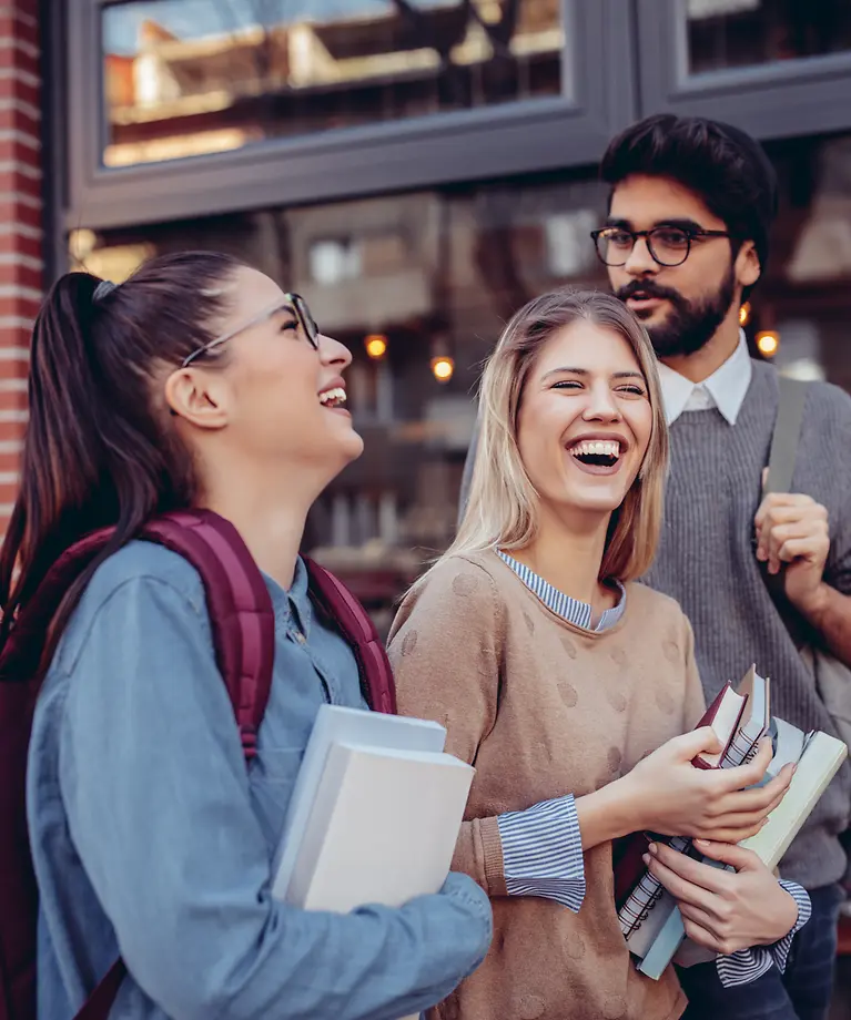 Drie studenten met boeken in hun handen lachen om. Op de achtergrond zie je een café.
